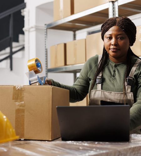 African american employee wearing industrial overall working in storehouse, preparing customers orders for delivery. Storage room employee checking shipping detalis on laptop computer, portrait