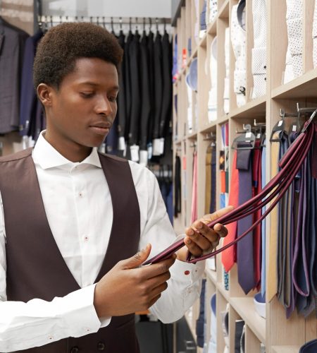 Handsome client of shop choosing necktie. African man holding red cravat looking at it. Young man wearing in white shirt and black waistcoat. Huge choice of clothing for men.